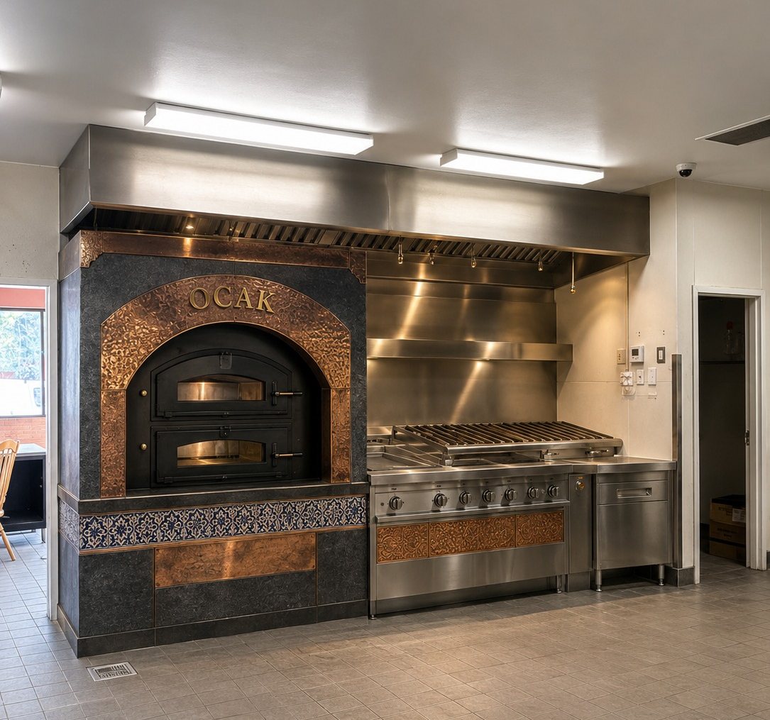 A copper-clad stone oven with the word OCAK worked into the metal, set above a band of blue and white tile, beside a commercial cooking line